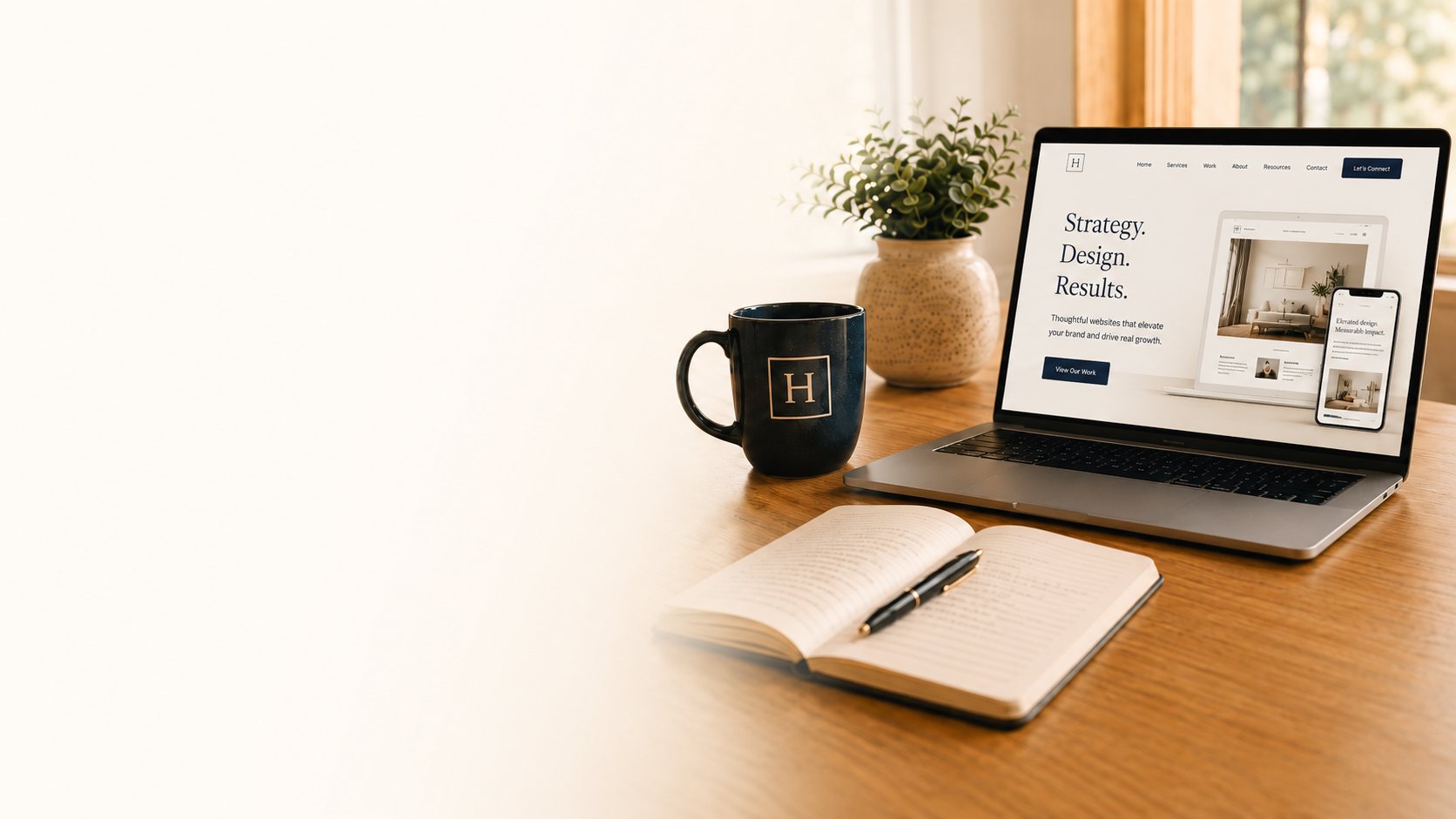 A laptop on a sunlit wooden desk showing a website design in progress, beside a Harper Studio mug and an open notebook